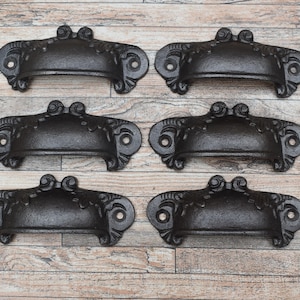 Peut inclure: Six poignées de tiroir en fonte noire avec des détails ornés. Les poignées sont disposées en deux rangées de trois sur une surface en bois.