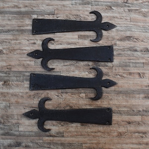 May include: Four black metal hinges with a fleur-de-lis design. The hinges are arranged in a vertical stack on a wooden background.