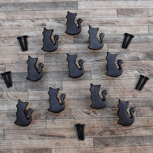 Puede incluir: Diez tiradores de cajón de metal negro en forma de gato con acabado cepillado, dispuestos en círculo sobre una superficie de madera. Se incluyen cuatro pares de tornillos.