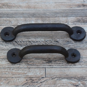 Könnte beinhalten: Zwei schwarze Schubladengriffe aus Gusseisen mit abgerundeten Enden. Die Griffe befinden sich auf einem Holzgrund.