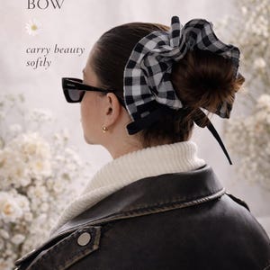 Puede incluir: Un accesorio para el cabello con lazo de cuadros blanco y negro. La imagen muestra a una persona que lleva el lazo, con el texto "GINGHAM BOW" y "carry beauty softly". La persona lleva una chaqueta de cuero negra y gafas de sol.