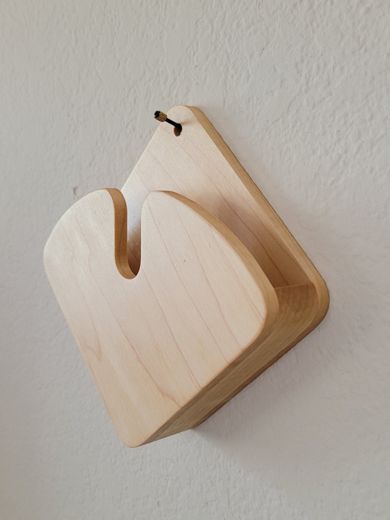 May include: A light-colored wooden wall-mounted organizer with a unique, curved design. The organizer has a diamond-shaped back with a U-shaped cutout and a small shelf. A small screw and black cord are attached at the top for hanging.