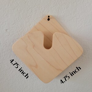 May include: A light-colored wooden wall-mounted guitar holder. The holder is diamond-shaped, measuring 4.75 inches on each side, with a U-shaped cutout to cradle the guitar neck. A small hole is drilled at the top for hanging.