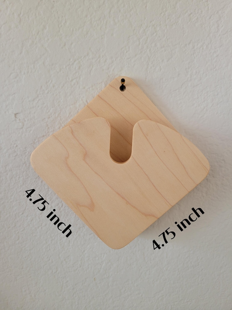 May include: A light-colored wooden wall-mounted guitar holder. The holder is diamond-shaped, measuring 4.75 inches on each side, with a U-shaped cutout to cradle the guitar neck. A small hole is drilled at the top for hanging.