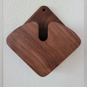 May include: A wall-mounted wooden organizer with a diamond-shaped design. The organizer is made of dark brown wood and features a curved opening in the center. A small hole is at the top for hanging. The wood grain is visible.