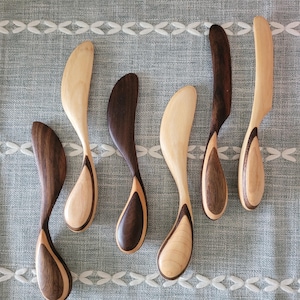 May include: Seven wooden butter knives with a unique, curved design. The knives are made of two different types of wood, creating a contrasting pattern. The knives are arranged in a row on a gray fabric background.