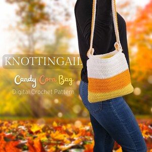 Puede incluir: Un bolso de crochet blanco, naranja y amarillo con un diseño de maíz dulce. El bolso se lleva como bolso de bandolera y tiene una correa larga. El bolso está delante de un fondo borroso de hojas de otoño.