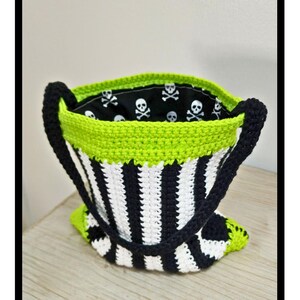 Puede incluir: Una bolsa de mano de crochet a rayas blancas y negras con ribete verde. La bolsa tiene un forro de tela con calaveras y huesos cruzados.