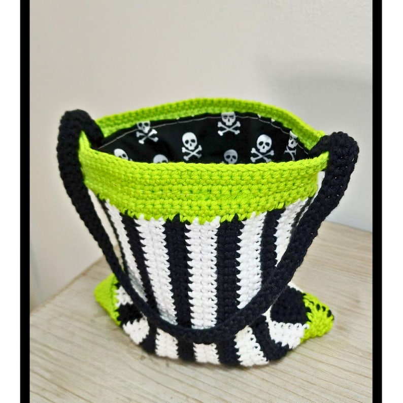 Puede incluir: Una bolsa de mano de crochet a rayas blancas y negras con ribete verde. La bolsa tiene un forro de tela con calaveras y huesos cruzados.