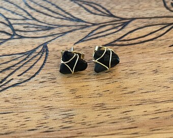 May include: A pair of black stone stud earrings. Each earring features a dark, irregularly shaped stone wrapped in gold-colored wire. The earrings are set against a wooden surface with a leaf-like pattern.