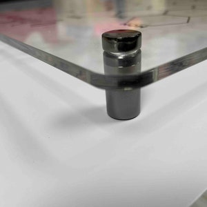 Pode incluir: Close-up de uma moldura de acr&iacute;lico transparente com um suporte de metal preto. O suporte tem uma parte superior redonda e uma base cil&iacute;ndrica.
