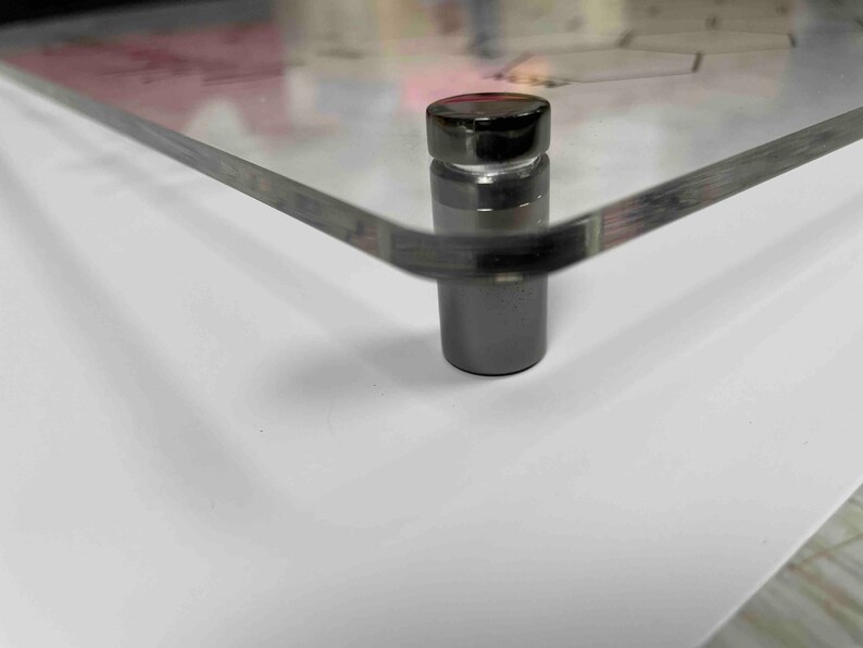 Pode incluir: Close-up de uma moldura de acr&iacute;lico transparente com um suporte de metal preto. O suporte tem uma parte superior redonda e uma base cil&iacute;ndrica.