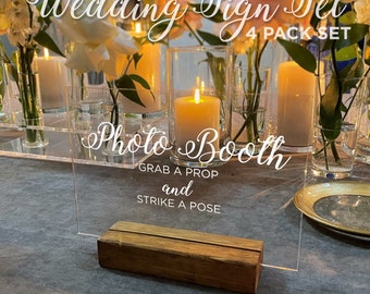 Op de afbeelding: Set van heldere acryl trouwtekens met de tekst "Wedding Sign Set 4 Pack Set" en "Photo Booth Grab a Prop and Strike a Pose". Het bord wordt vastgehouden door een houten voet, met kaarsen en bloemen op de achtergrond.
