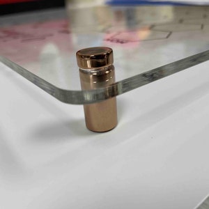 Pode incluir: Close-up de um suporte de metal dourado-rosado com uma superf&iacute;cie de vidro transparente. O suporte &eacute; usado para exibir obras de arte ou fotos.