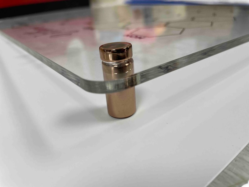 Pode incluir: Close-up de um suporte de metal dourado-rosado com uma superf&iacute;cie de vidro transparente. O suporte &eacute; usado para exibir obras de arte ou fotos.