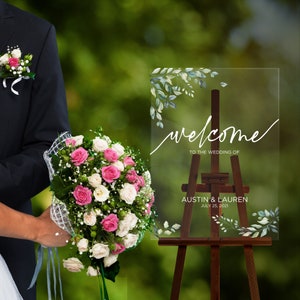 May include: A clear acrylic wedding welcome sign with a leafy design and the text "welcome to the wedding of Austin & Lauren July 25, 2021" on a wooden easel.
