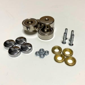 Könnte beinhalten: Eine Auswahl an Hardware-Komponenten, darunter silberfarbene Magnete, Schrauben, Muttern und goldfarbene Unterlegscheiben. Die Artikel sind auf einer weißen Oberfläche angeordnet und zeigen ihre metallischen Oberflächen und verschiedenen Formen.