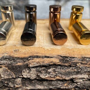 Pode incluir: Um conjunto de oito rolhas de garrafa de metal em v&aacute;rios acabamentos, incluindo prata, preto, ouro e ouro rosa. As rolhas est&atilde;o dispostas em uma fila sobre uma superf&iacute;cie de madeira.