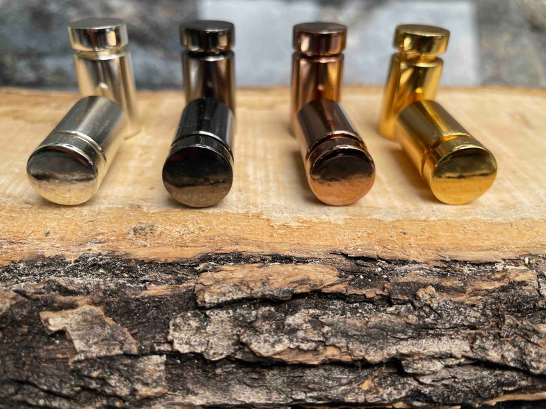 Pode incluir: Um conjunto de oito rolhas de garrafa de metal em v&aacute;rios acabamentos, incluindo prata, preto, ouro e ouro rosa. As rolhas est&atilde;o dispostas em uma fila sobre uma superf&iacute;cie de madeira.