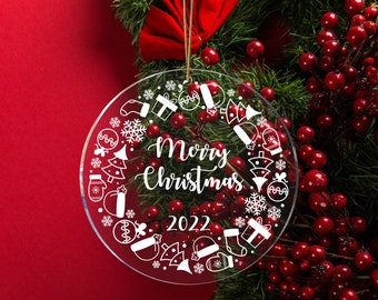 May include: A clear, circular Christmas decoration with the text "Merry Christmas 2022" in white script. The ornament features festive symbols such as snowflakes, presents, and mittens. It hangs from a brown string against a red backdrop.