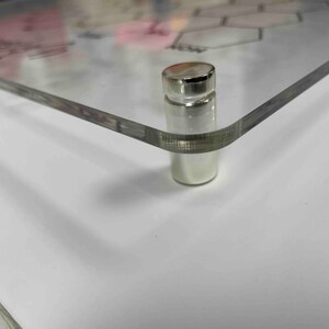Pode incluir: Close-up de um suporte de metal prateado com uma parte superior lisa e arredondada. O suporte est&aacute; preso a uma superf&iacute;cie de acr&iacute;lico transparente.