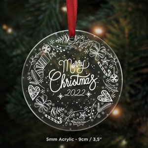 May include: A clear acrylic Christmas bauble with a white design of a wreath with snowflakes, candy canes, holly, and the text "Merry Christmas 2022". The bauble is 9cm in diameter and is made of 5mm acrylic.