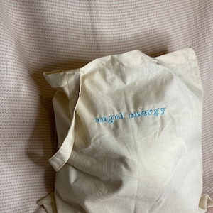 以下が含まれることがあります： 薄いクリーム色のトートバッグ。水色の刺繍で「angel energy」と書かれています。バッグは柔らかい生地でできており、持ち手が付いています。背景はピンクと白の模様の生地です。