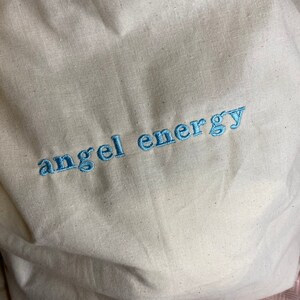 以下が含まれることがあります： クリーム色のトートバッグ。 軽めの青い糸で「angel energy」の文字が刺繍されています。 バッグは、おそらく綿またはリネンなどの天然素材で作られており、シンプルでカジュアルなデザインです。