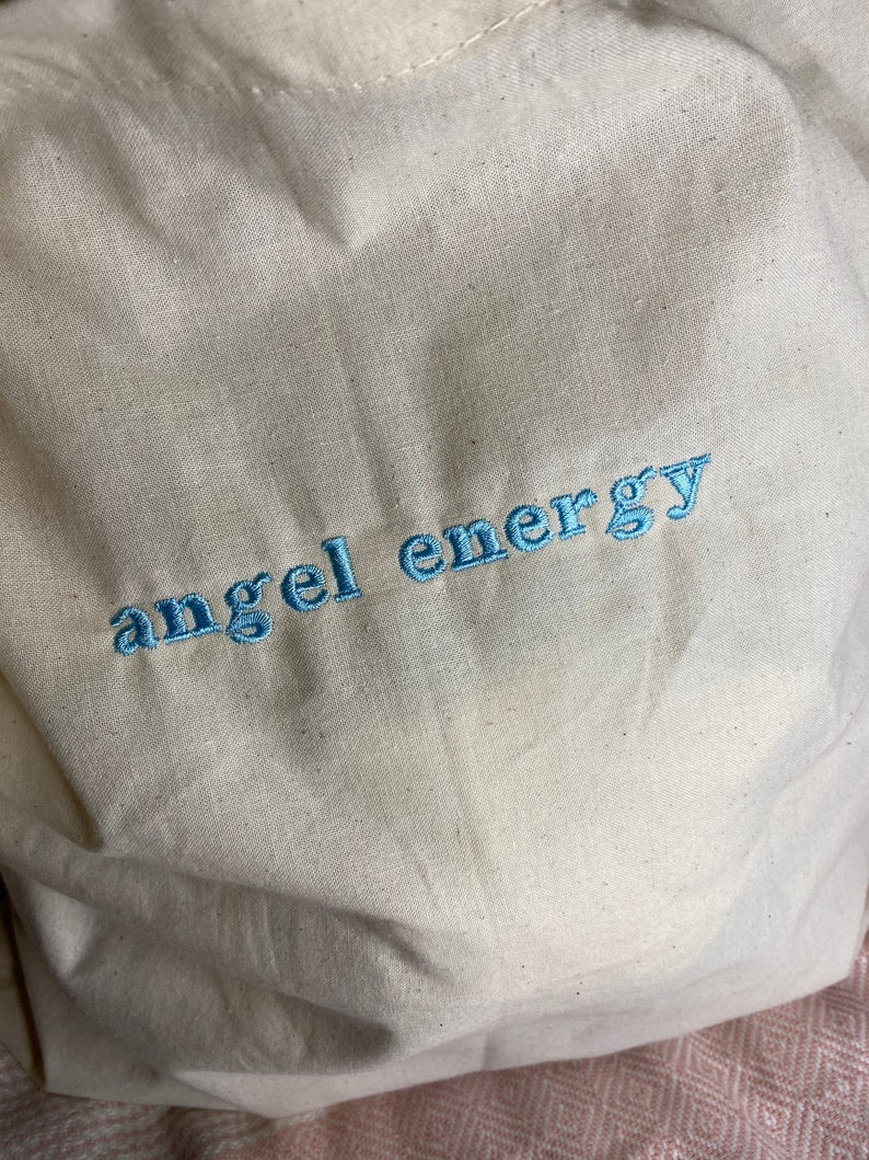 以下が含まれることがあります： クリーム色のトートバッグ。 軽めの青い糸で「angel energy」の文字が刺繍されています。 バッグは、おそらく綿またはリネンなどの天然素材で作られており、シンプルでカジュアルなデザインです。