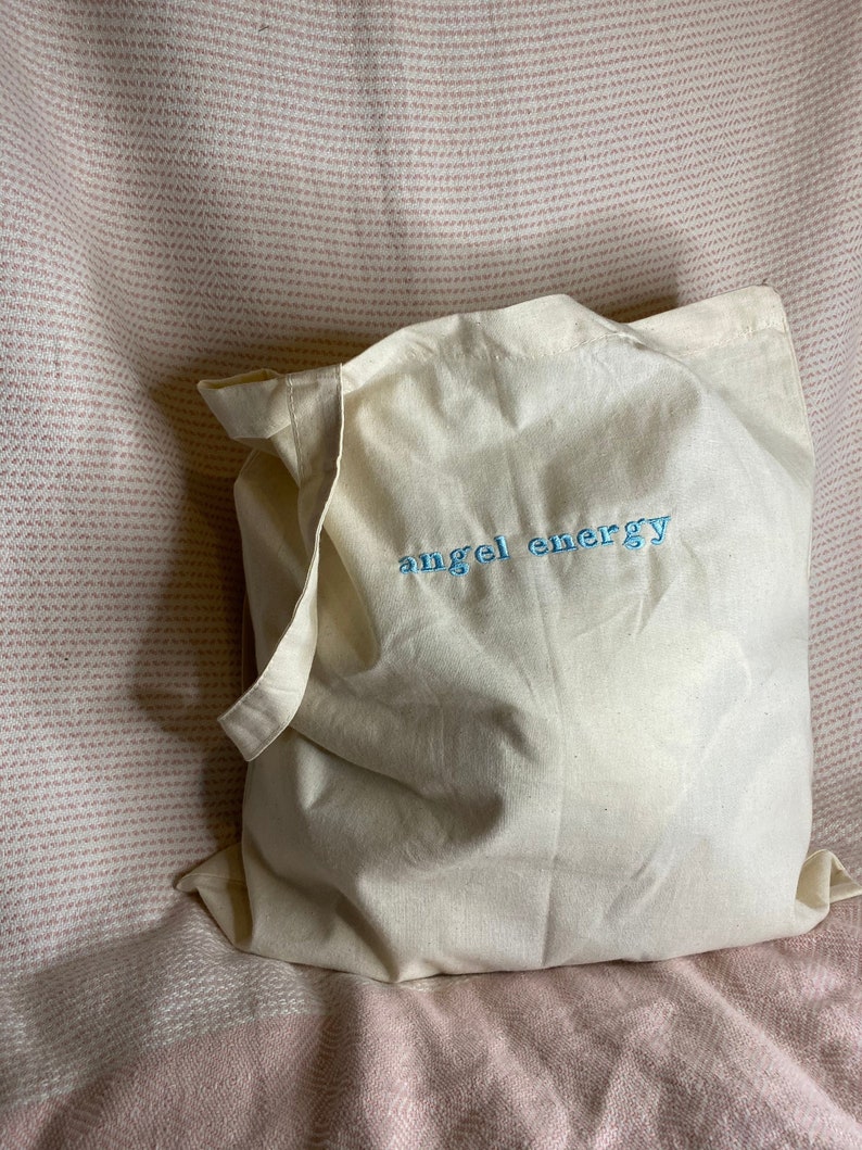 以下が含まれることがあります： 薄いクリーム色のトートバッグ。水色の刺繍で「angel energy」と書かれています。バッグは柔らかい生地でできており、持ち手が付いています。背景はピンクと白の模様の生地です。