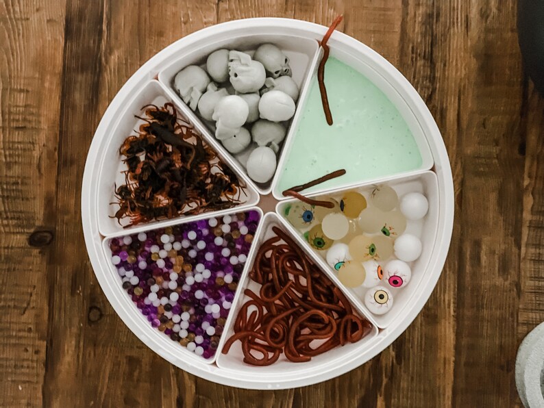 May include: A white circular tray divided into six sections, each filled with different Halloween-themed items. The sections contain plastic skulls, fake spiders, small purple and white beads, red gummy worms, green slime, and plastic eyeballs.