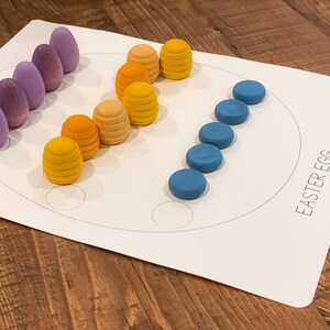 K&ouml;nnte beinhalten: Ein wei&szlig;es Brett mit einem darauf gezeichneten Kreis. Auf dem Brett steht "Easter Egg" gedruckt. Um den Kreis herum sind violette, blaue und gelbe Holzeier angeordnet.