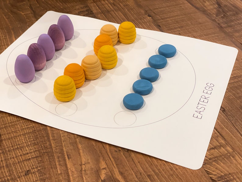 K&ouml;nnte beinhalten: Ein wei&szlig;es Brett mit einem darauf gezeichneten Kreis. Auf dem Brett steht "Easter Egg" gedruckt. Um den Kreis herum sind violette, blaue und gelbe Holzeier angeordnet.