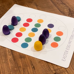 K&ouml;nnte beinhalten: Ein wei&szlig;es Brett mit dem Text "Easter Egg Pattern Completion" auf der rechten unteren Ecke. Das Brett hat einen Kreis mit 12 leeren Kreisen und 12 farbigen Kreisen in einem Muster. Es befinden sich 6 lilafarbene Holzeier und 1 gelbes Holzei auf dem Brett.