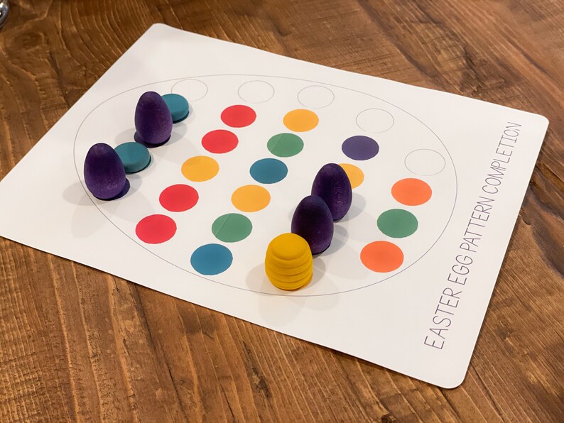 K&ouml;nnte beinhalten: Ein wei&szlig;es Brett mit dem Text "Easter Egg Pattern Completion" auf der rechten unteren Ecke. Das Brett hat einen Kreis mit 12 leeren Kreisen und 12 farbigen Kreisen in einem Muster. Es befinden sich 6 lilafarbene Holzeier und 1 gelbes Holzei auf dem Brett.