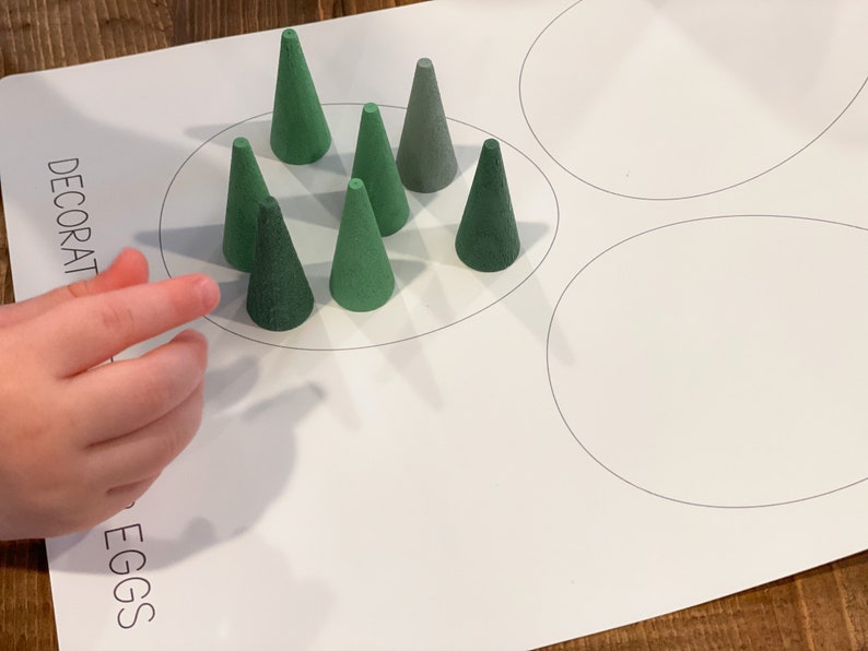 K&ouml;nnte beinhalten: Ein wei&szlig;es Blatt Papier mit den Worten "DECORATE EGGS" darauf gedruckt. Auf dem Papier sind drei Kreise gezeichnet, und in einem der Kreise ist eine Gruppe von neun gr&uuml;nen kegelf&ouml;rmigen Objekten angeordnet.