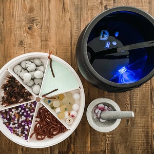 May include: A white plastic tray with six compartments filled with Halloween-themed candy and treats. The compartments contain gummy worms, fake eyeballs, small plastic skulls, purple and white beads, brown and red candy, and green slime. The tray is on a wooden table next to a black cauldron with a blue light inside.