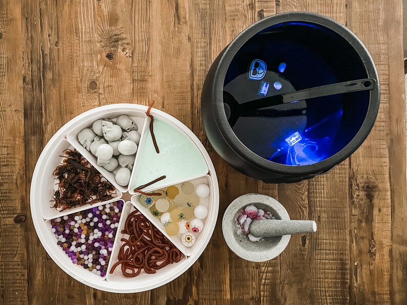 May include: A white plastic tray with six compartments filled with Halloween-themed candy and treats. The compartments contain gummy worms, fake eyeballs, small plastic skulls, purple and white beads, brown and red candy, and green slime. The tray is on a wooden table next to a black cauldron with a blue light inside.