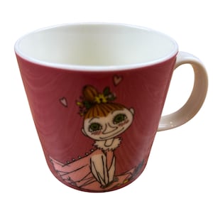 May include: A ceramic mug with a red exterior and a white interior. The mug features a cartoon illustration of a character with green eyes, a pink dress, and a floral hairpiece. The handle is white.