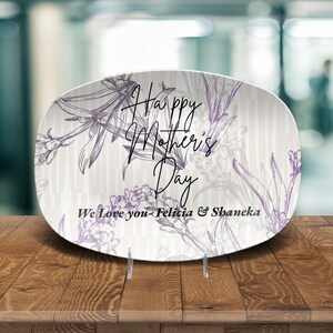 May include: Oval decorative plate with a floral design in purple and gray. The plate has the text "Happy Mother's Day" and "We Love you-Felicia & Shaneka". The plate is on a clear stand.
