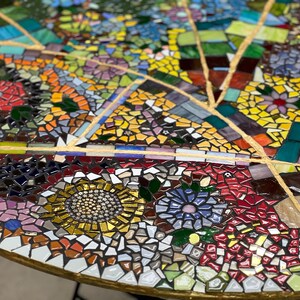 May include: A circular mosaic table top with a colorful design featuring flowers, leaves, and geometric shapes. The mosaic is made of small, irregular pieces of glass and tile in shades of blue, green, yellow, red, and white.