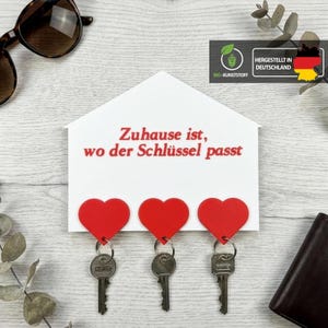 Può includere: Un portachiavi a forma di casa bianca con tre portachiavi rossi a forma di cuore. Il portachiavi ha il testo tedesco "Zuhause ist, wo der Schlüssel passt" in rosso. Occhiali da sole e un portafoglio sullo sfondo.