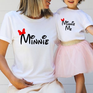 Könnte beinhalten: Zwei weiße T-Shirts mit einer roten Schleife und dem Text "Minnie" in Schwarz. Das größere Shirt sagt "Minnie" und das kleinere Shirt sagt "My Minnie Me".