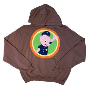 May include: A brown hooded sweatshirt with a large, colorful patch featuring a cartoon pig wearing a police officer's hat and uniform. The pig is waving and has a surprised expression.