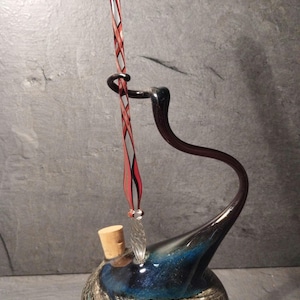 Könnte beinhalten: Eine Glaskaraffe mit rundem Boden und wirbelnden blauen, schwarzen und silbernen Mustern, verschlossen mit einem Korken. Ein langer, schlanker Glasstab mit rotem und klarem gedrehtem Design ruht in einem geschwungenen schwarzen Glashalter.