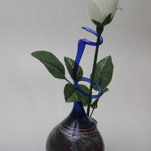 Könnte beinhalten: Eine einzelne weiße Rose mit grünen Blättern in einer blauen und braunen Glasvase mit einem geschwungenen blauen Glasstiel.