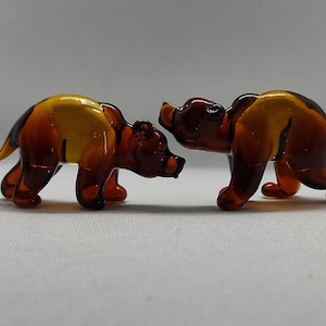 Może przedstawiać: Dwie małe szklane figurki niedźwiedzi, jedna brązowa, a druga bursztynowa z żółtym paskiem, stojące naprzeciwko siebie.