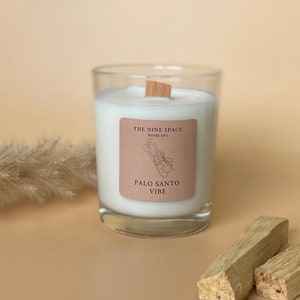 Könnte beinhalten: Eine weiße Kerze in einem Glasgefäß mit einem Holzdeckel. Das Etikett auf dem Gefäß lautet "The Nine Space Home Spa Palo Santo Vibe".