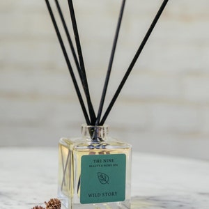 Könnte beinhalten: Eine klare Glasflasche mit schwarzen Diffusorstäbchen und einem grünen Etikett mit der Aufschrift "THE NINE BEAUTY & HOME SPA WILD STORY".