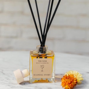 Puede incluir: Una botella de vidrio transparente con un líquido marrón y palitos difusores negros. La botella tiene una etiqueta que dice "The Nine Beauty & Home Spa" y "Scandal Story".